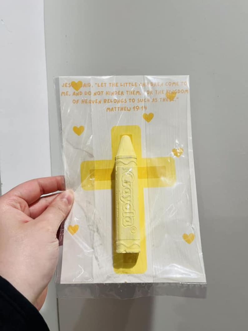 May include: A yellow crayon with the word "Crayola" printed on it, packaged with a white card featuring a yellow cross and hearts. The card includes a Bible verse from Matthew 19:14, "Let the little children come to me, and do not hinder them, for the kingdom of heaven belongs to such as these."