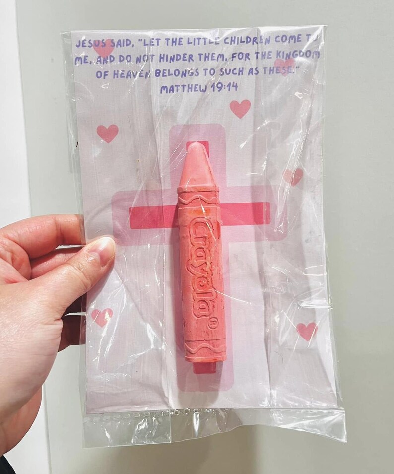 May include: A pink crayon with the word "Crayola" printed on it, packaged in a clear plastic bag with a white card. The card has a pink cross and two pink hearts. The card also has the text "JESUS SAID, "LET THE LITTLE CHILDREN COME TO ME, AND DO NOT HINDER THEM, FOR THE KINGDOM OF HEAVEN BELONGS TO SUCH AS THESE." MATTHEW 19:14"