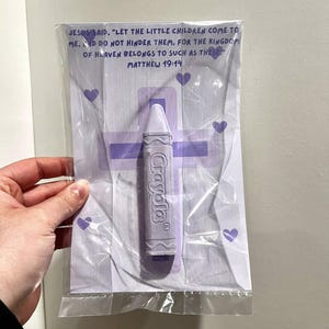 May include: A purple crayon with the word "Crayola" printed on it, packaged in a clear plastic bag with a purple cross and hearts. The bag also includes a Bible verse: "Jesus said, 'Let the little children come to me. And do not hinder them, for the kingdom of heaven belongs to such as these.' Matthew 19:14"