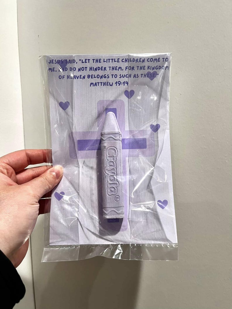 May include: A purple crayon with the word "Crayola" printed on it, packaged in a clear plastic bag with a purple cross and hearts. The bag also includes a Bible verse: "Jesus said, 'Let the little children come to me. And do not hinder them, for the kingdom of heaven belongs to such as these.' Matthew 19:14"