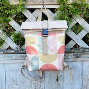 May include: A beige and patterned backpack with a geometric design of colorful semi-circles. The backpack has beige straps, a black buckle, and a beige flap. The bag is hanging on a hook against a wooden fence.