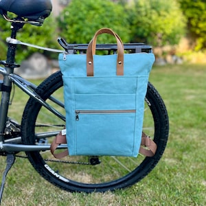 May include: A light blue canvas backpack with brown leather handles and straps. The bag features a horizontal zippered front pocket, a top zipper closure, and two vertical leather handle attachments. It is mounted on a black bicycle rear rack.
