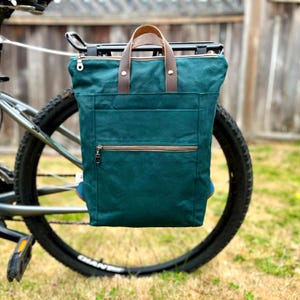 May include: A dark teal canvas bicycle pannier bag featuring brown leather top handles, a horizontal zippered front pocket, and a main top zipper closure. The bag is mounted onto a black metal rear bicycle rack with silver hardware accents.