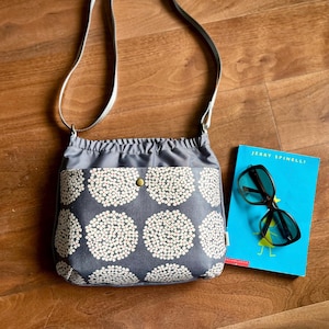 May include: Gray crossbody bag with a white floral pattern on the front pocket. The bag has a drawstring closure and a silver strap. A blue book and black sunglasses are next to the bag.