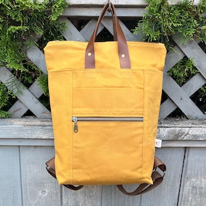 May include: Mustard yellow backpack with brown leather handles and straps. The backpack has a front zipped pocket and a top handle. The bag is hanging on a wooden fence with green foliage in the background.