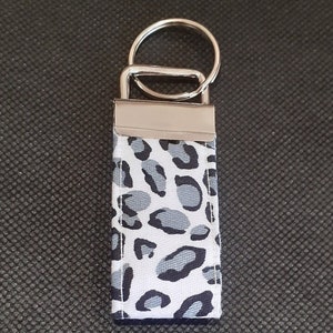 May include: A rectangular keychain with a silver metal ring and clasp. The fabric section displays a white background with a grey and black leopard print pattern. The keychain is designed for holding keys.