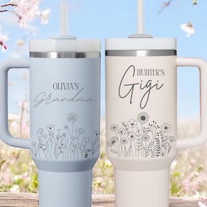 Pode incluir: Dois copos isolados personalizados com al&ccedil;as e palhinhas. Um &eacute; azul claro com "Olivia's Grandma" e design floral. O outro &eacute; branco sujo com "Hunter's Gigi" e design floral. Ambos t&ecirc;m tampas prateadas.