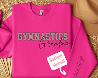 May include: Pink sweatshirt with the text "Gymnastics Grandma" in gray and black lettering. The right sleeve has the text "Gymnastics Grandma" in black lettering. A white speech bubble with the text "Custom Sleeve!" is on the front of the sweatshirt.