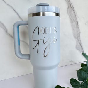 Pode incluir: Um copo t&eacute;rmico azul claro com uma al&ccedil;a e uma borda prateada. O copo tem as palavras "ODIN'S Gigi" em uma fonte prateada. A tampa &eacute; cinza claro e a al&ccedil;a &eacute; azul claro.