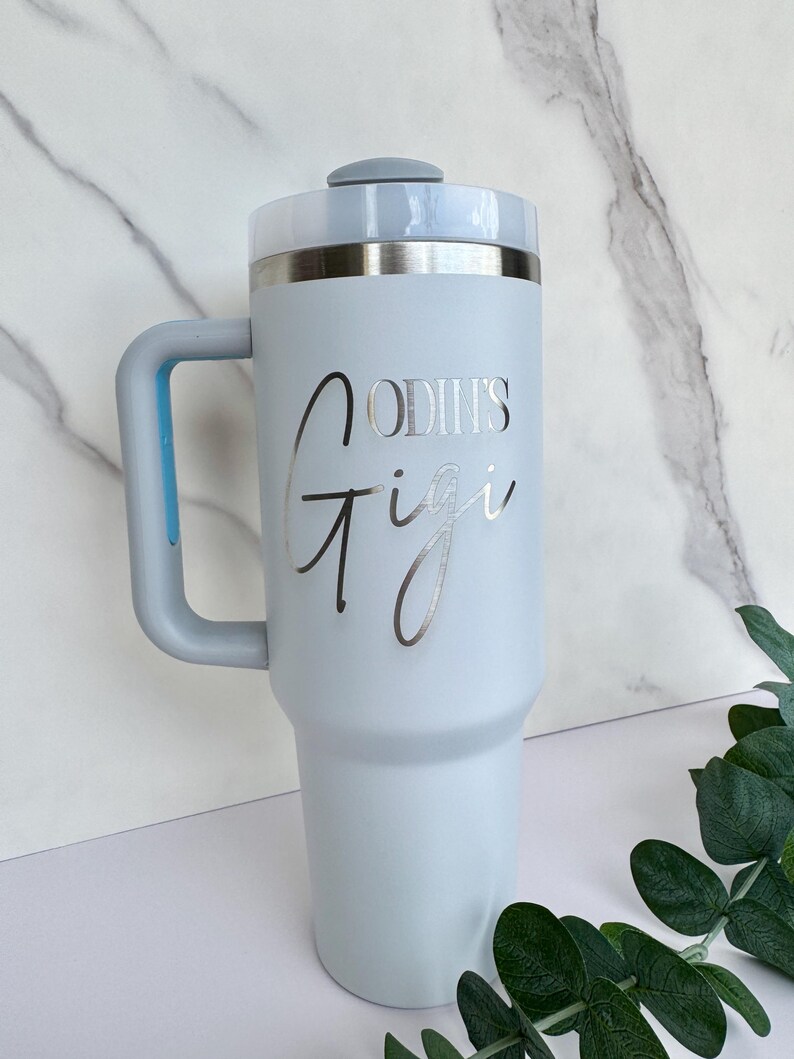 Pode incluir: Um copo t&eacute;rmico azul claro com uma al&ccedil;a e uma borda prateada. O copo tem as palavras "ODIN'S Gigi" em uma fonte prateada. A tampa &eacute; cinza claro e a al&ccedil;a &eacute; azul claro.