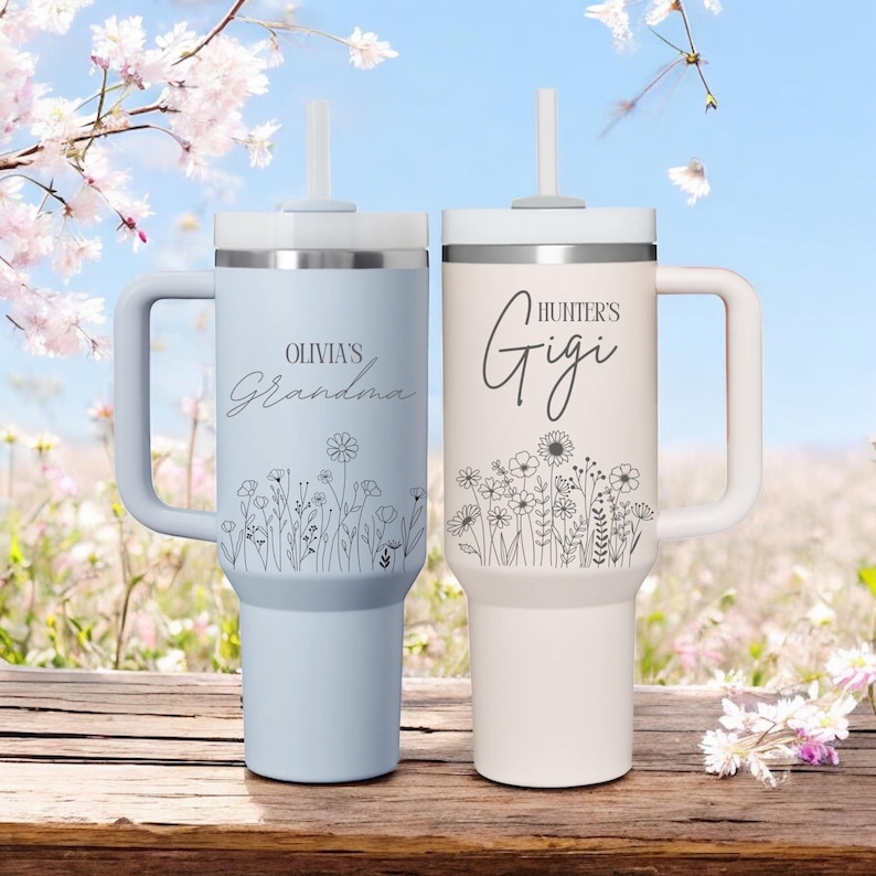 Pode incluir: Dois copos isolados personalizados com al&ccedil;as e palhinhas. Um &eacute; azul claro com "Olivia's Grandma" e design floral. O outro &eacute; branco sujo com "Hunter's Gigi" e design floral. Ambos t&ecirc;m tampas prateadas.