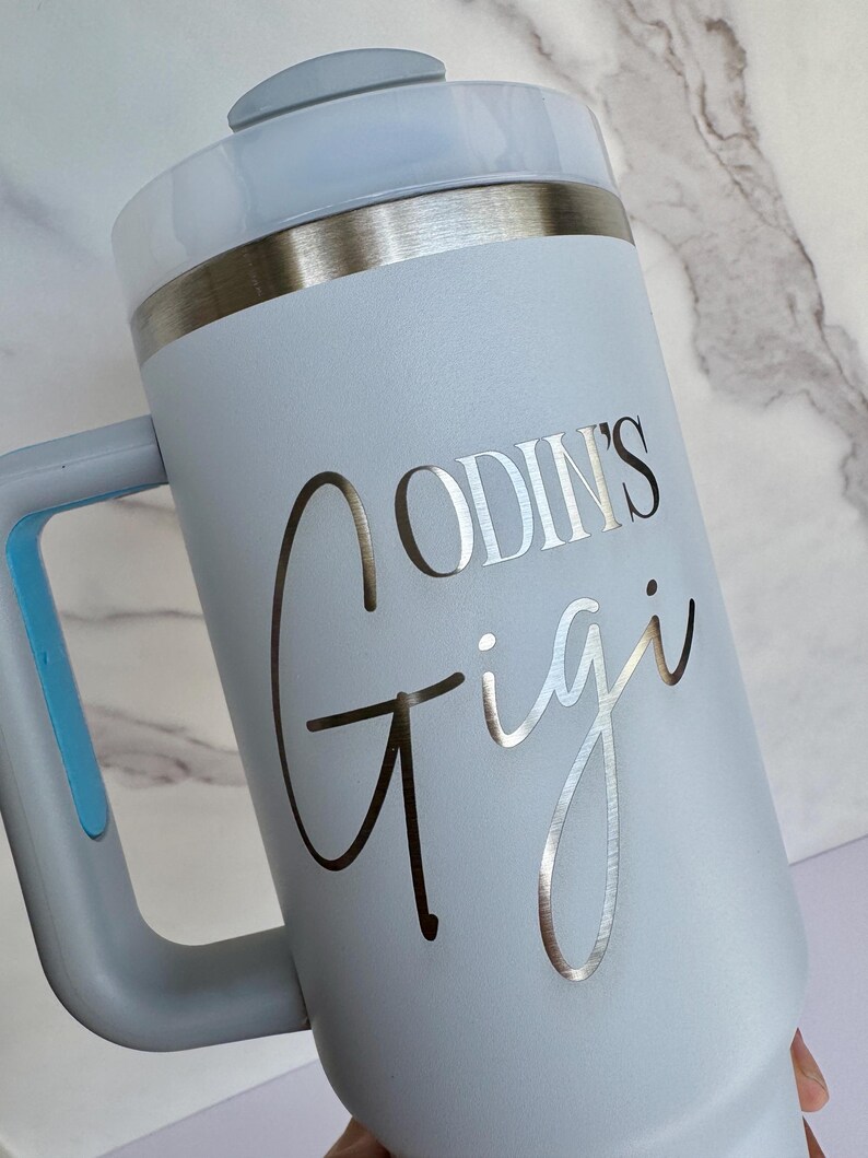Pode incluir: Um copo t&eacute;rmico azul claro com uma al&ccedil;a e uma borda prateada. O copo apresenta o texto "ODIN'S Gigi" em prata. A tampa &eacute; azul clara com um bot&atilde;o cinza.