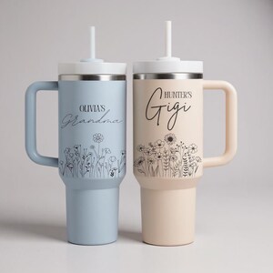 Pode incluir: Duas canecas isoladas com al&ccedil;as e canudos. Uma &eacute; azul clara com "Olivia's Grandma" e design floral. A outra &eacute; bege com "Hunter's Gigi" e design floral. Ambas t&ecirc;m tampas brancas.