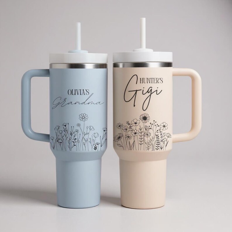 Pode incluir: Duas canecas isoladas com al&ccedil;as e canudos. Uma &eacute; azul clara com "Olivia's Grandma" e design floral. A outra &eacute; bege com "Hunter's Gigi" e design floral. Ambas t&ecirc;m tampas brancas.