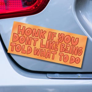 May include: A light orange bumper sticker with the text "HONK IF YOU DON'T LIKE BEING TOLD WHAT TO DO" in a rounded, bubble-like font.