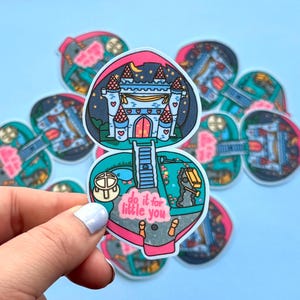 Puede incluir: Una pegatina en forma de coraz&oacute;n con un dise&ntilde;o de castillo bajo un cielo nocturno, con el texto "do it for little you". La pegatina tiene un borde rosa y turquesa. La pegatina est&aacute; sujeta por una mano sobre un fondo azul.