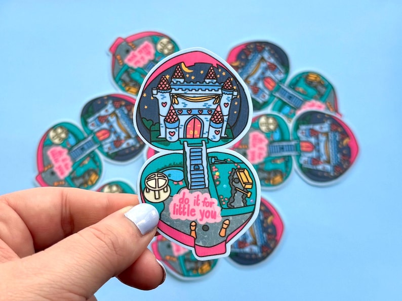 Puede incluir: Una pegatina en forma de coraz&oacute;n con un dise&ntilde;o de castillo bajo un cielo nocturno, con el texto "do it for little you". La pegatina tiene un borde rosa y turquesa. La pegatina est&aacute; sujeta por una mano sobre un fondo azul.