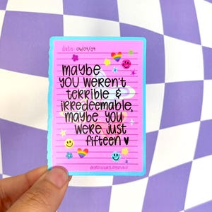 May include: A pink sticker with the text "maybe YOU weren't terrible & irredeemable, maybe you were just fifteen" in black. The sticker has a light blue border and is decorated with smiley faces, hearts, and rainbows.