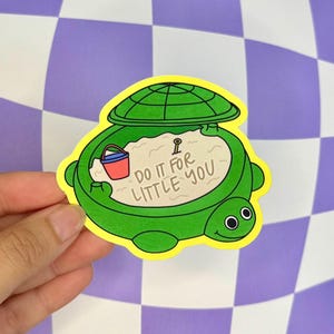 May include: A green turtle-shaped sticker with a yellow border. The turtle has a sand-filled shell with a small red and blue bucket. Text on the sticker reads "DO IT FOR LITTLE YOU."