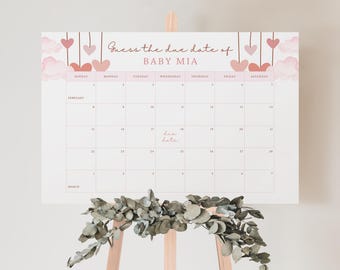 May include: A white calendar sign with the text Guess the due date of BABY MIA, featuring pink heart and cloud illustrations, hanging strings, and a grid layout for February and March, displayed on a wooden easel with green foliage.