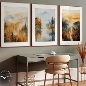 Könnte beinhalten: Drei gerahmte Aquarelle, die neblige Waldszenen mit goldenen Farbtönen darstellen. Die Bilder hängen an einer grauen Wand über einem Schreibtisch mit einem Stuhl.