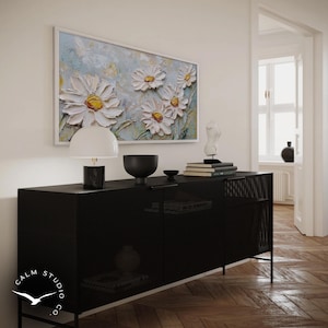 May include: A black cabinet with a perforated design, topped with a white lamp, a black bowl, and books. Above, a framed painting of white daisies with yellow centers hangs on the wall. The painting is set against a light blue background.