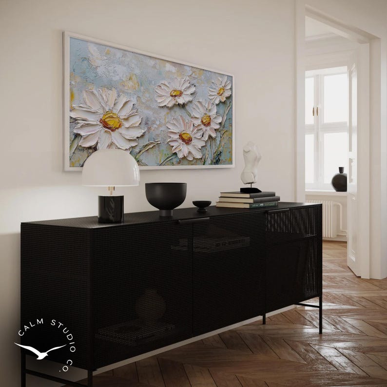 May include: A black cabinet with a perforated design, topped with a white lamp, a black bowl, and books. Above, a framed painting of white daisies with yellow centers hangs on the wall. The painting is set against a light blue background.