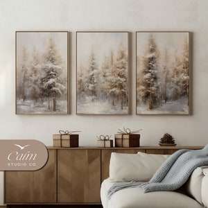 May include: Three framed canvas prints of a winter forest scene with snow-covered trees in shades of brown and white. The prints are hung on a white wall above a wooden cabinet and a white couch with a gray throw blanket.