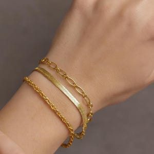 May include: Three gold bracelets of different designs are stacked on a wrist. The bracelets include a chain link, a herringbone, and a delicate rope chain. The jewelry is displayed against a neutral background.