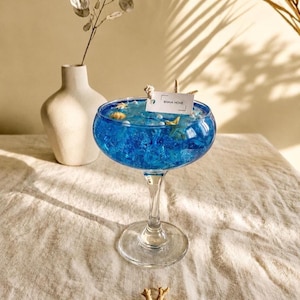 Peut inclure: Une bougie en gel bleu dans une coupe en verre transparent, remplie de petites perles blanches, de coquillages et d'une étoile de mer. Une étiquette rectangulaire blanche avec le texte BHAVA HOME est fixée à la mèche. Posée sur un tissu texturé.