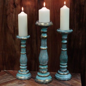 May include: Set of three teal blue candle holders with gold accents. The candle holders are made of wood and have a distressed finish. Each holder has a round base and a tall, slender column.