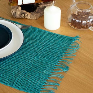 May include: A teal burlap placemat with a fringed edge, on a wooden table. A white candle, a plate with a dark blue rim, and a glass teapot are also visible. The placemat adds a rustic touch to the table setting.