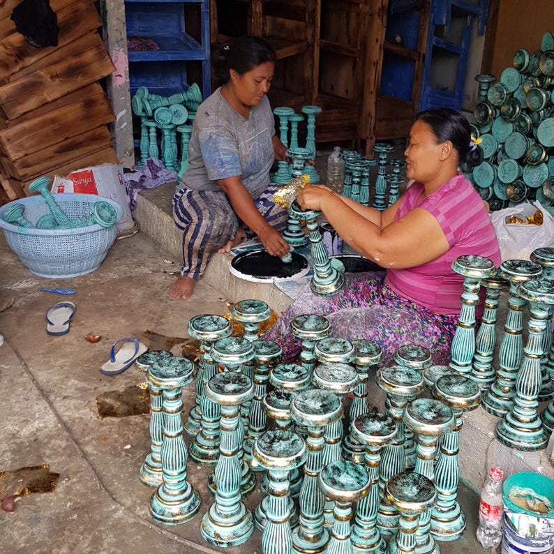 May include: Two people are working at a table, painting wooden candle holders a teal colour. The candle holders are stacked and ready to be painted.
