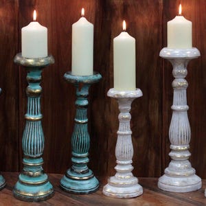 May include: Set of six candle holders, three in white with gold accents and three in teal with gold accents. The candle holders are made of wood and have a distressed finish. Each holder has a round base and a tall, slender stem.