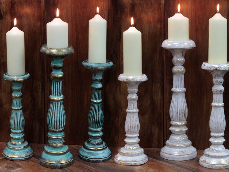 May include: Set of six candle holders, three in white with gold accents and three in teal with gold accents. The candle holders are made of wood and have a distressed finish. Each holder has a round base and a tall, slender stem.