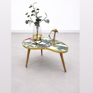 Puede incluir: Una mesa de centro con forma de riñón, borde de metal dorado y tres patas de madera cónicas. La superficie superior es blanca con un estampado botánico de hojas verdes y flores rojas. Incluye un jarrón de cristal y una figura de ave de metal.