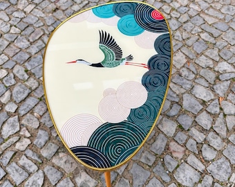 Könnte beinhalten: Ein nierenförmiger Beistelltisch mit cremefarbener Tischplatte, die eine fliegende Kranich-Illustration und konzentrische Kreismuster in Petrol, Dunkelblau und Rosa zeigt. Die Platte ist mit einem dünnen goldfarbenen Metallrand eingefasst.