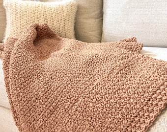Könnte beinhalten: Eine weiche, braune Strickdecke, die über ein weißes Sofa mit einem weißen Kissen gelegt wird.