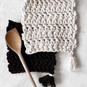 May include: Two macrame pot holders, one black and one cream, with a wooden spoon. The pot holders have a woven texture and a tassel at the corner. The wooden spoon is light brown and has a long handle.