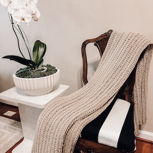 May include: A white orchid plant in a white ceramic pot sits on a white marble pedestal table next to a wooden chair with a black and white striped cushion. A beige knitted throw blanket is draped over the chair.