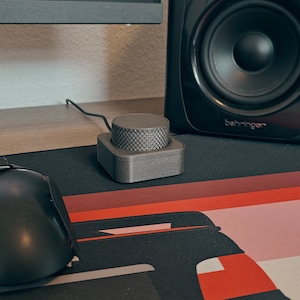 Może przedstawiać: Czarna mysz komputerowa i szare pokrętło regulacji głośności leżą na wzorzystej podkładce pod mysz. Czarny głośnik znajduje się w tle. Pokrętło regulacji głośności jest połączone z przewodem.