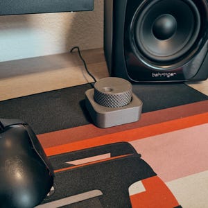 Puede incluir: Un mando de control de volumen gris con una parte superior texturizada se encuentra sobre una alfombrilla de ratón con estampado negro y rojo. También se ven un ratón de ordenador negro y un altavoz negro. El altavoz tiene la marca "behringer".