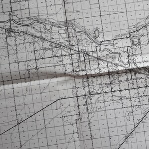 May include: A black and white map with a grid pattern and a river winding through the center. The map is folded in half, and the fold line is visible.