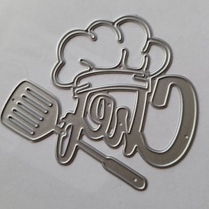 May include: A silver-colored metal die cut in the shape of the word "Chef" with a chef's hat and spatula. The word "Chef" is written in a stylized script font. The spatula is on the left side of the word.