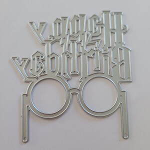 May include: A silver metal die cut in the shape of glasses with the words "You're a wizard, Harry" cut out above the lenses. The die cut is for crafting and scrapbooking projects.