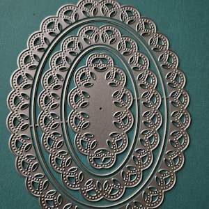 May include: A set of three nested oval-shaped metal die cuts. Each die cut features a decorative, scalloped edge with a repeating pattern of small dots and leaf-like shapes. The dies are silver-colored and sit on a teal background.