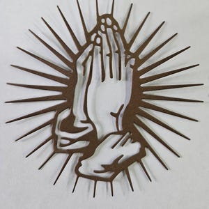 Op de afbeelding: Een bruin metalen wandkunstwerk met handen in gebed, omringd door een zonnesterontwerp. Het kunstwerk is uit metaal gesneden en heeft een gedetailleerd silhouet, geschikt voor religieuze of spirituele decoratie.