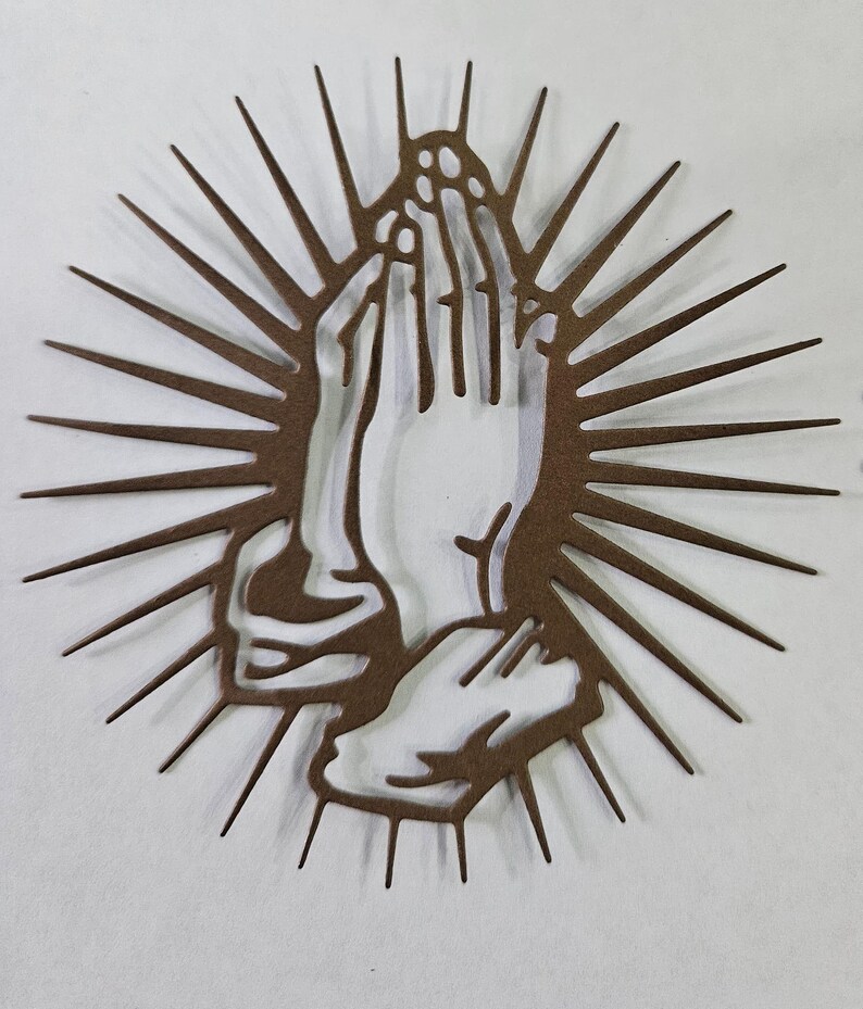 Op de afbeelding: Een bruin metalen wandkunstwerk met handen in gebed, omringd door een zonnesterontwerp. Het kunstwerk is uit metaal gesneden en heeft een gedetailleerd silhouet, geschikt voor religieuze of spirituele decoratie.