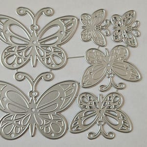 May include: A collection of die-cut metal butterfly shapes in various sizes. The butterflies have intricate wing designs with cut-out patterns and delicate antennae. The shapes are connected by thin metal strips, suggesting they are part of a crafting set.