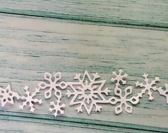 May include: A collection of white snowflake cutouts in various sizes and designs, arranged in a horizontal line. The snowflakes are placed against a light blue and green wooden background, creating a wintery theme.